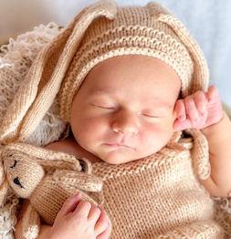 Sleeping newborn baby in beige knitted outfit and hat, holding a small knitted bunny toy